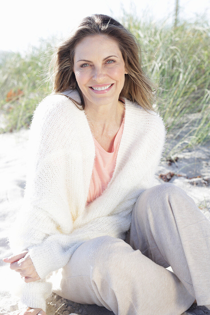 A long-haired woman sitting in the sand on the beach wearing a light jumper and trousers