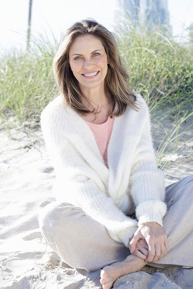 A long-haired woman sitting in the sand on the beach wearing a light jumper and trousers