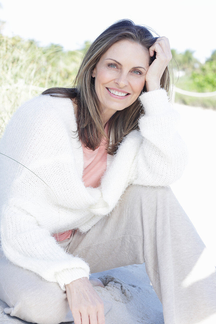 A long-haired woman sitting in the sand on the beach wearing a light jumper and trousers