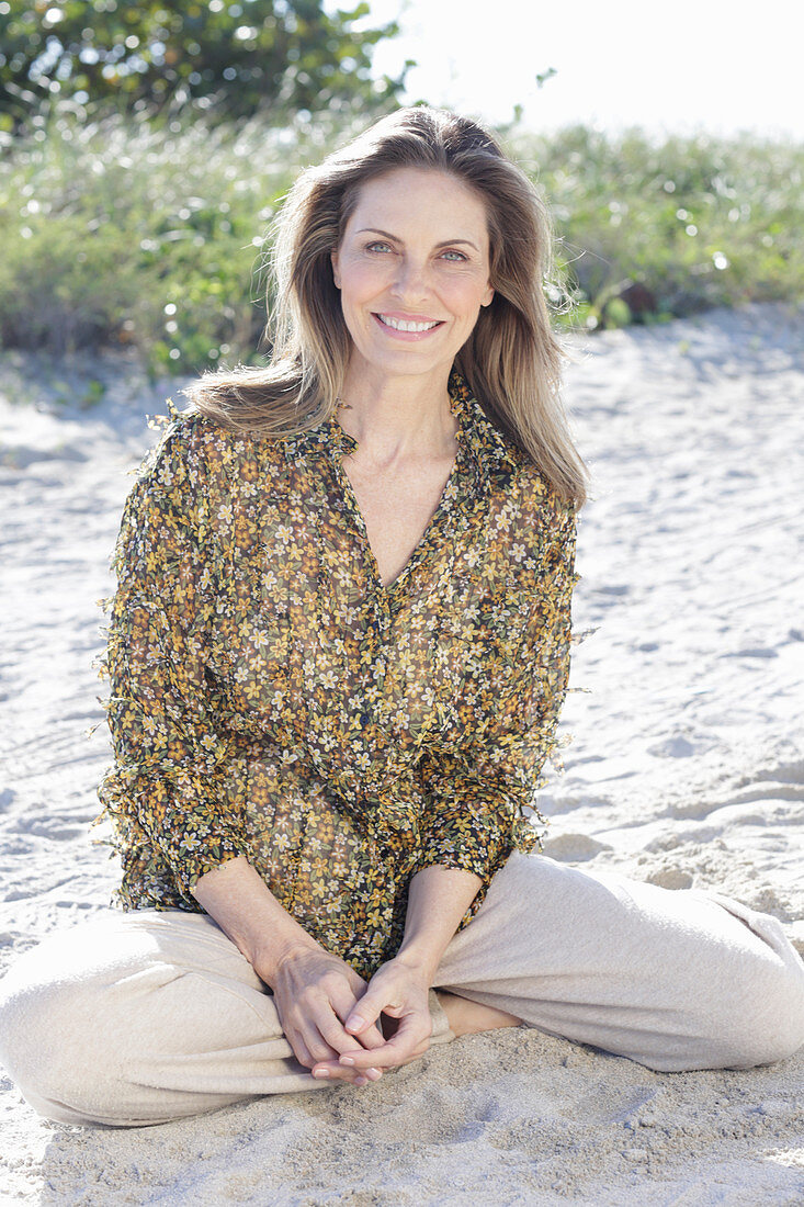 A long-haired woman sitting in the sand wearing a floral shirt and light trousers