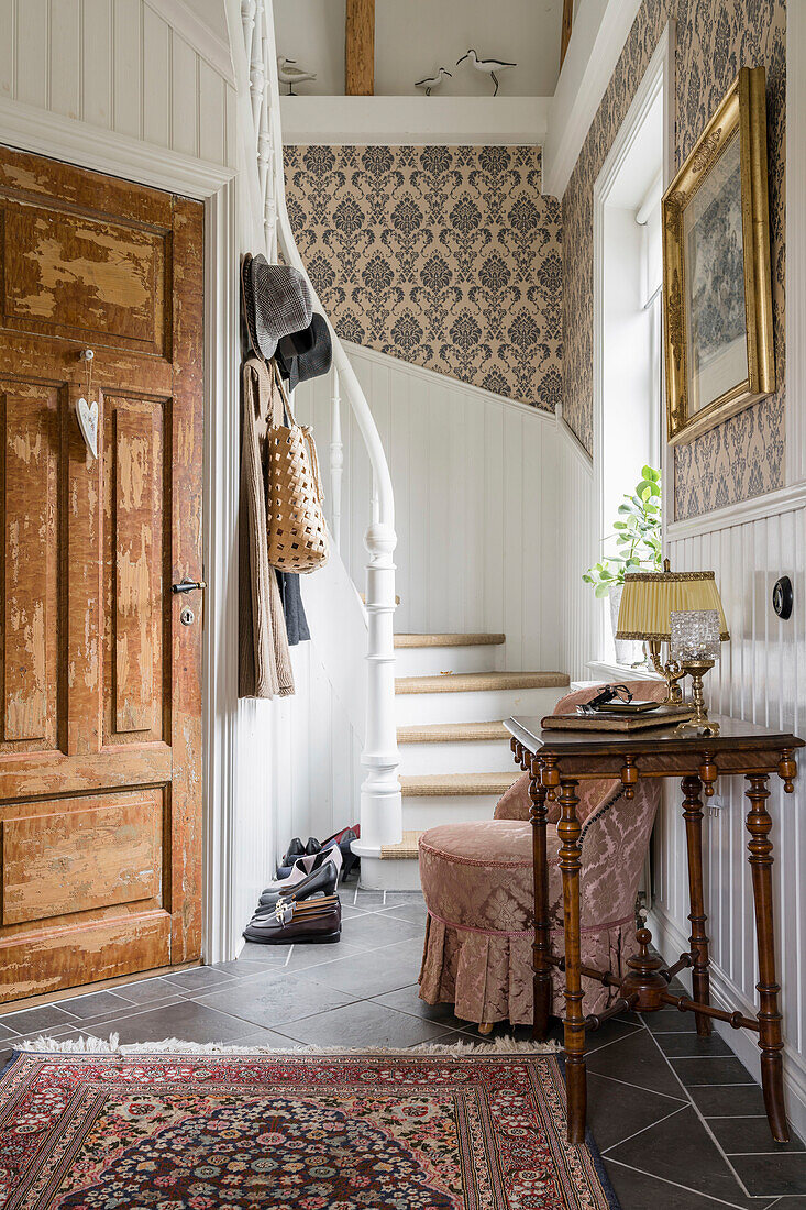 Antique console table and easy chair in hall with white wood panelling and medallion wallpaper