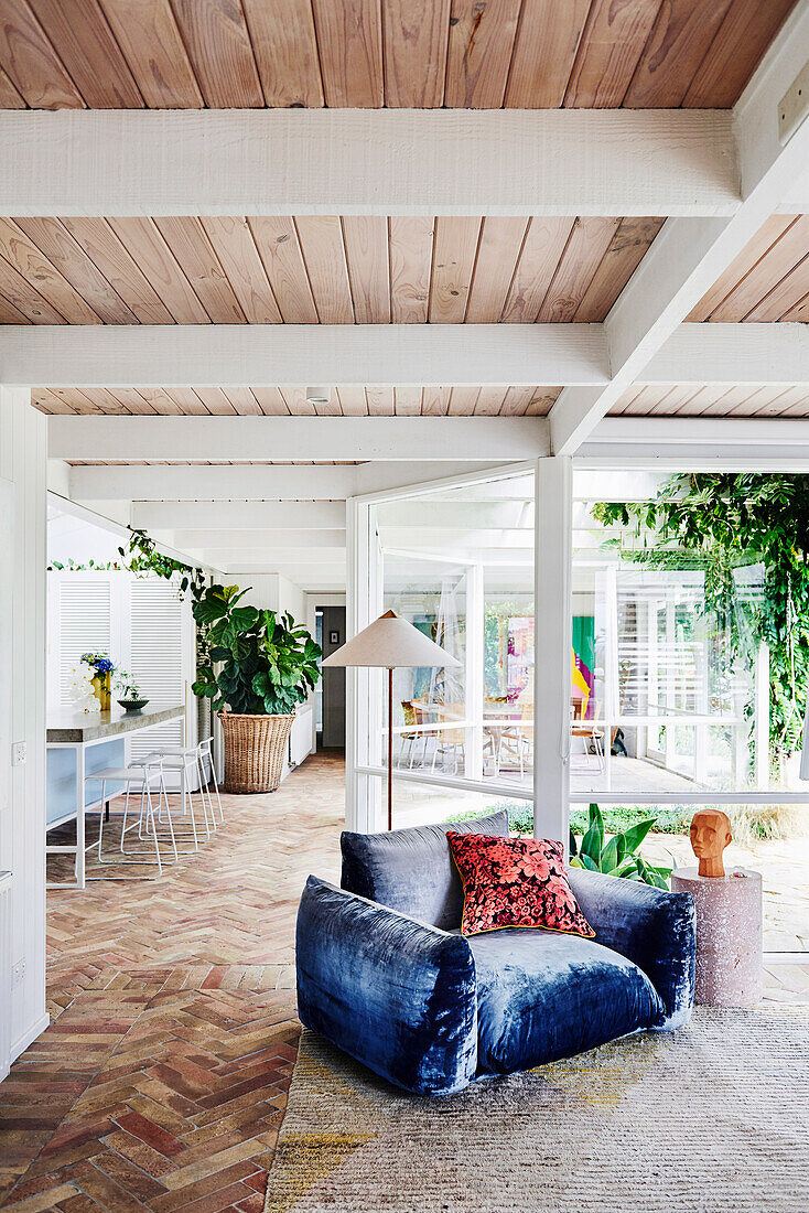 Armchair with blue velvet upholstery in front of floor-to-ceiling glazing in open living room with wooden ceiling