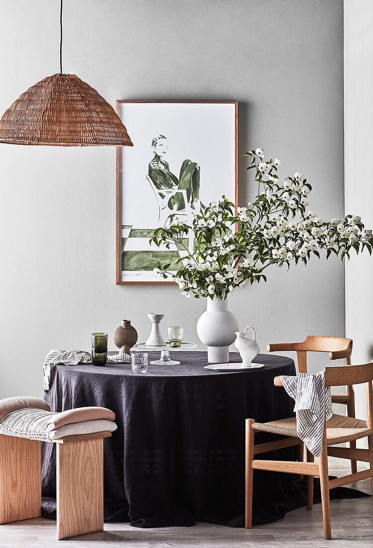 Vase with blossom branches on round table with purple tablecloth, stools and chairs, modern art on the wall