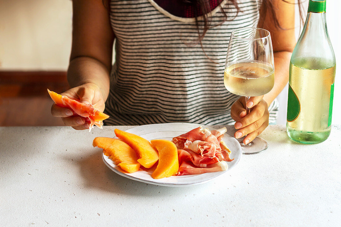 Female holding a slice of cantaloupe wrapped in prosciutto and a glass of wine