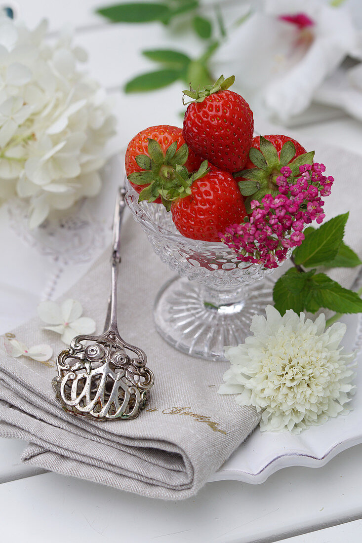Stillleben mit Erdbeeren in Glasschälchen, Blüten und silberner Gebäckzange