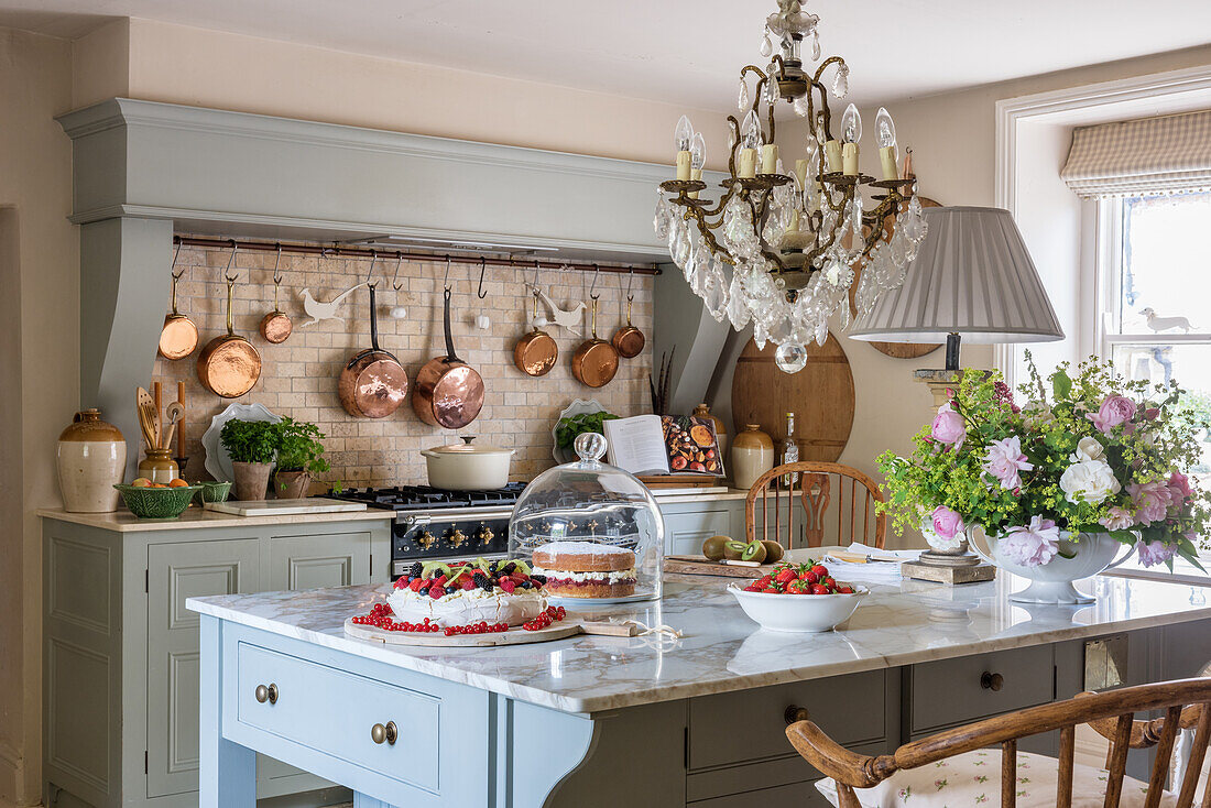 Cakes on kitchen table with marble worktop below French chandelier; copper pans on wall