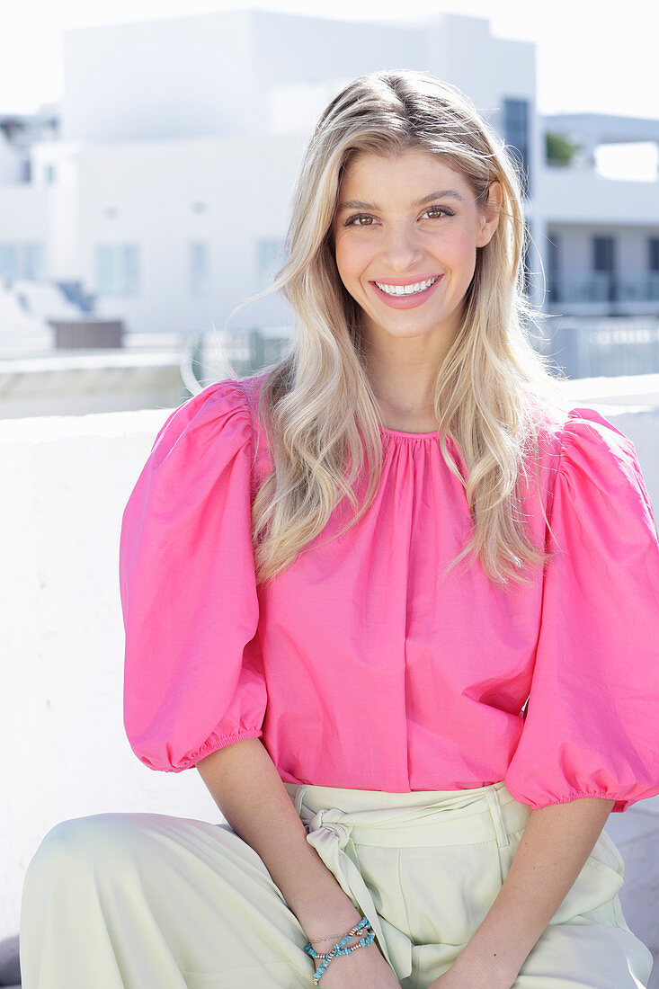 A young blonde woman wearing a pink top with puffed sleeves and white summer trousers