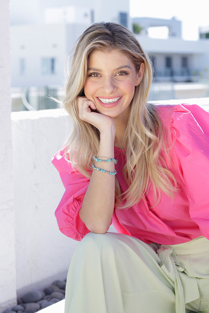 A young blonde woman wearing a pink top with puffed sleeves and white summer trousers