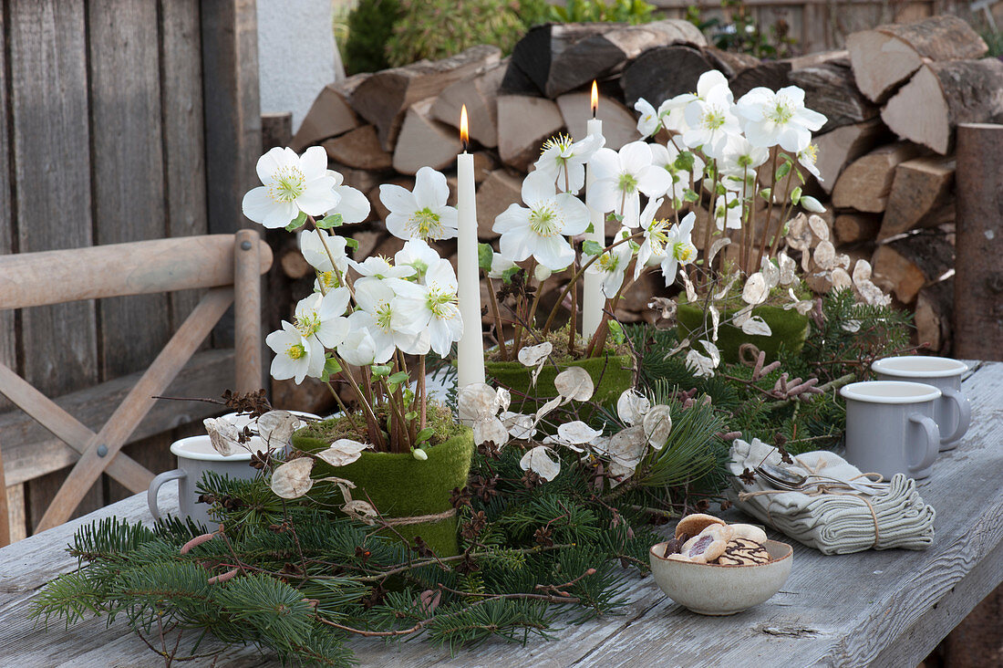 Christmas table decorations on the terrace: candles, Christmas roses in a felt pot, fir branches and silver coins, stacks of firewood in the background