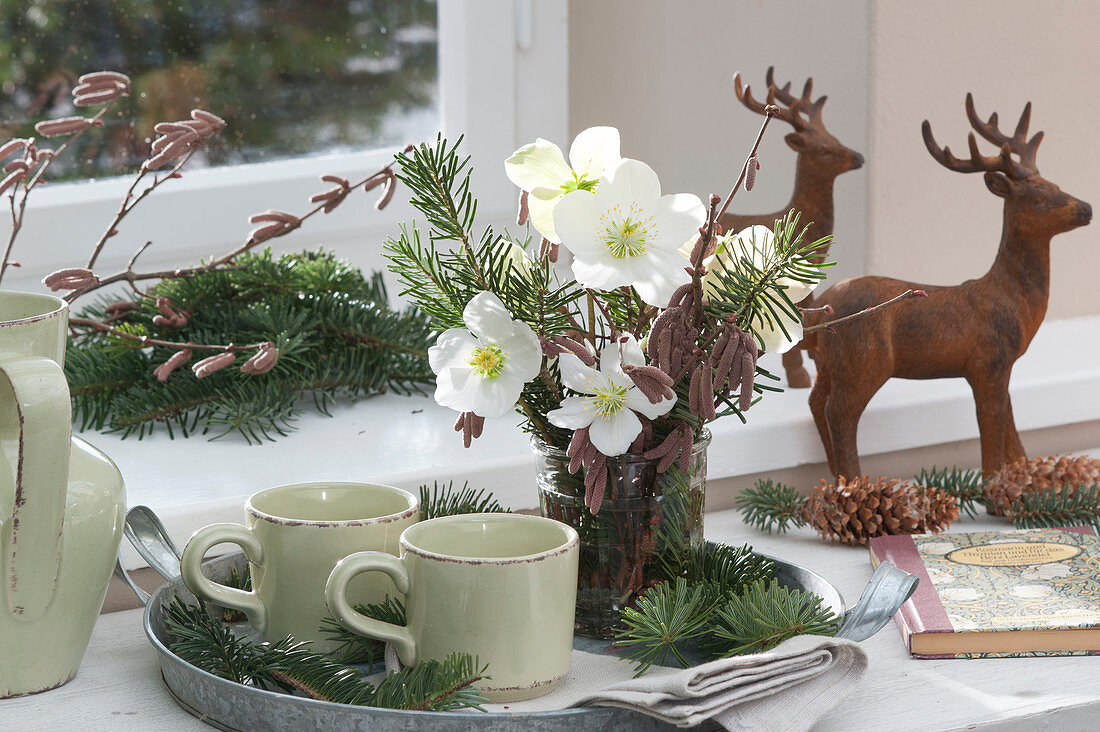 Small Christmas bouquet of Christmas roses, fir branches, and hazel branches on a tray with mugs, deer figures, and pinecones as decoration