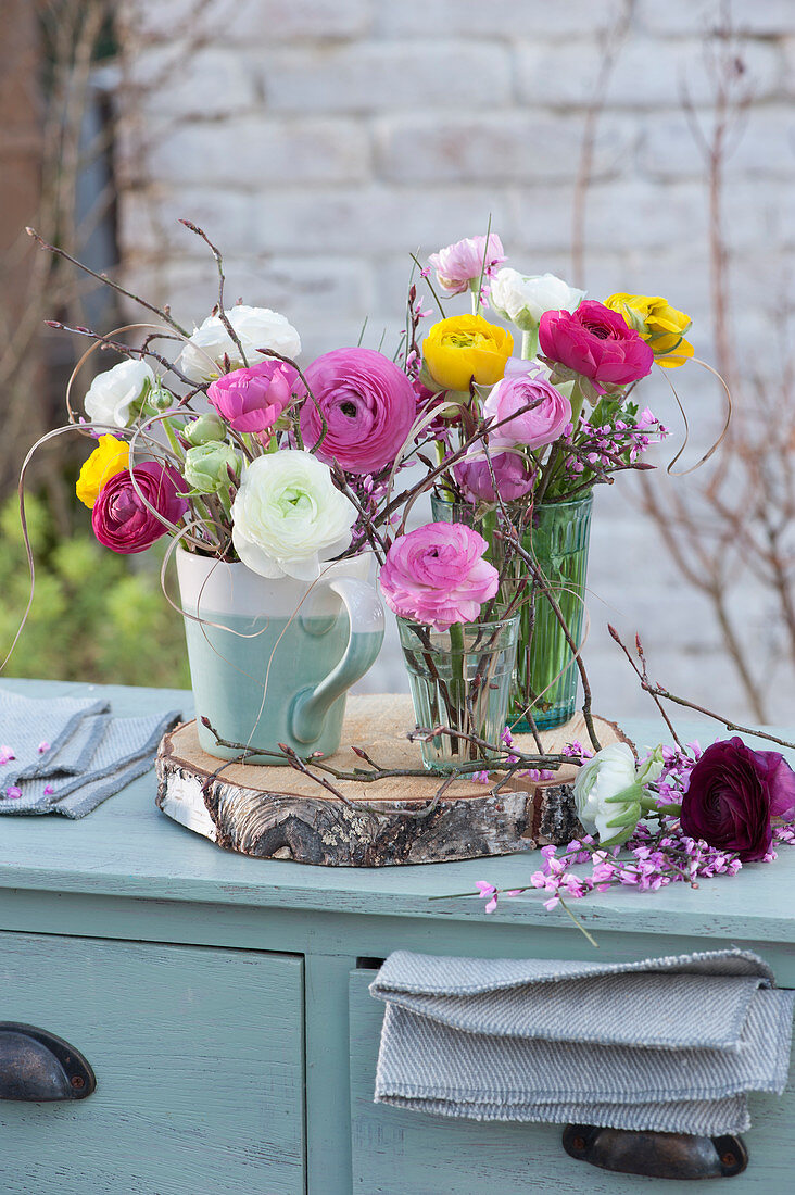 Small bouquets of ranunculus, broom, and twigs in cup and glasses on a wooden disc