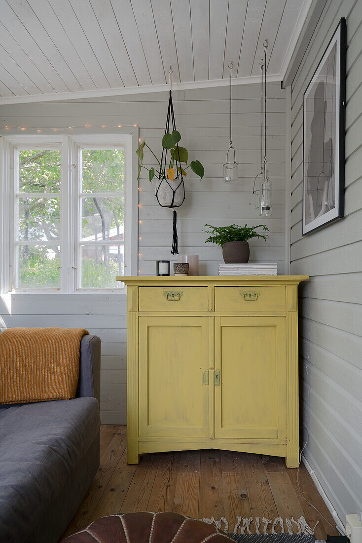 Yellow chest of drawers in the corner of a room, above it macramé flower hanging plants and lanterns
