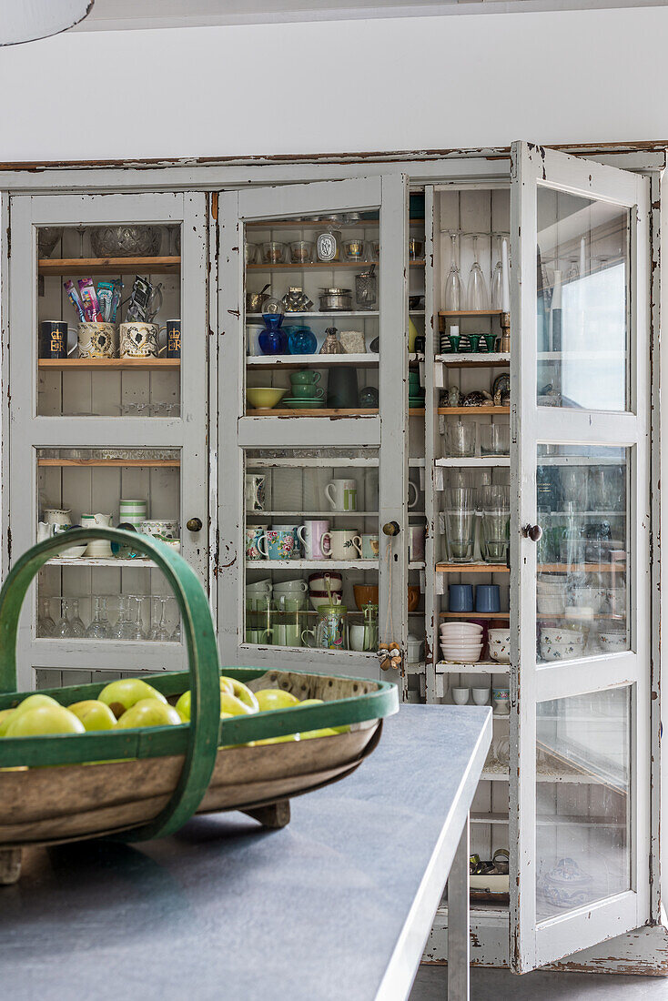 Antique crockery in display cabinet and basket of apples on kitchen island in foreground