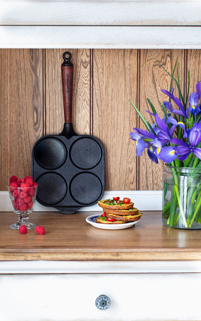 Pancake-Pfanne, Glas mit Himbeeren, Pancake-Stapel und Iris-Strauß auf Küchenablage
