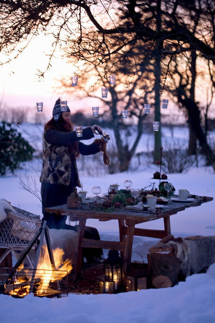 A woman hanging lanterns on a tree above a table laid in a snowy garden