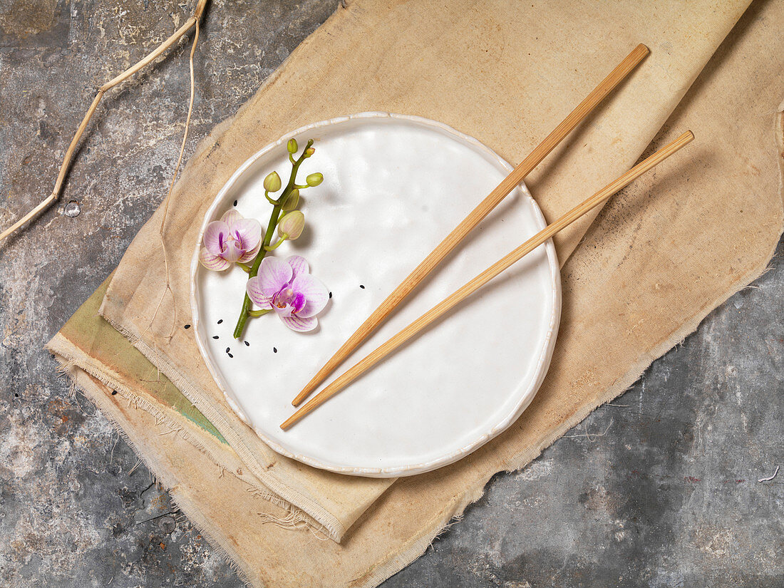 Orchideenblütenzweig und Essstäbchen auf weißem Teller und Leinentuch