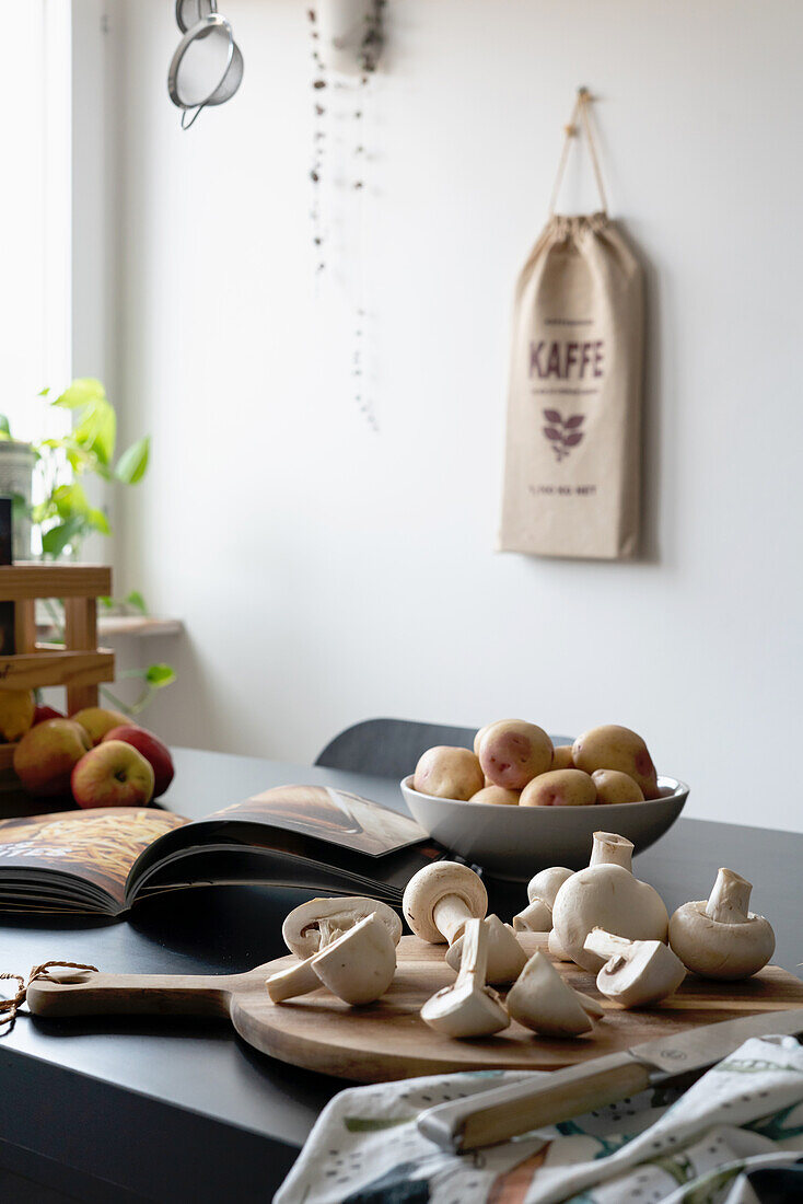 Wooden cutting board with mushrooms, potatoes, and cookbook on kitchen island