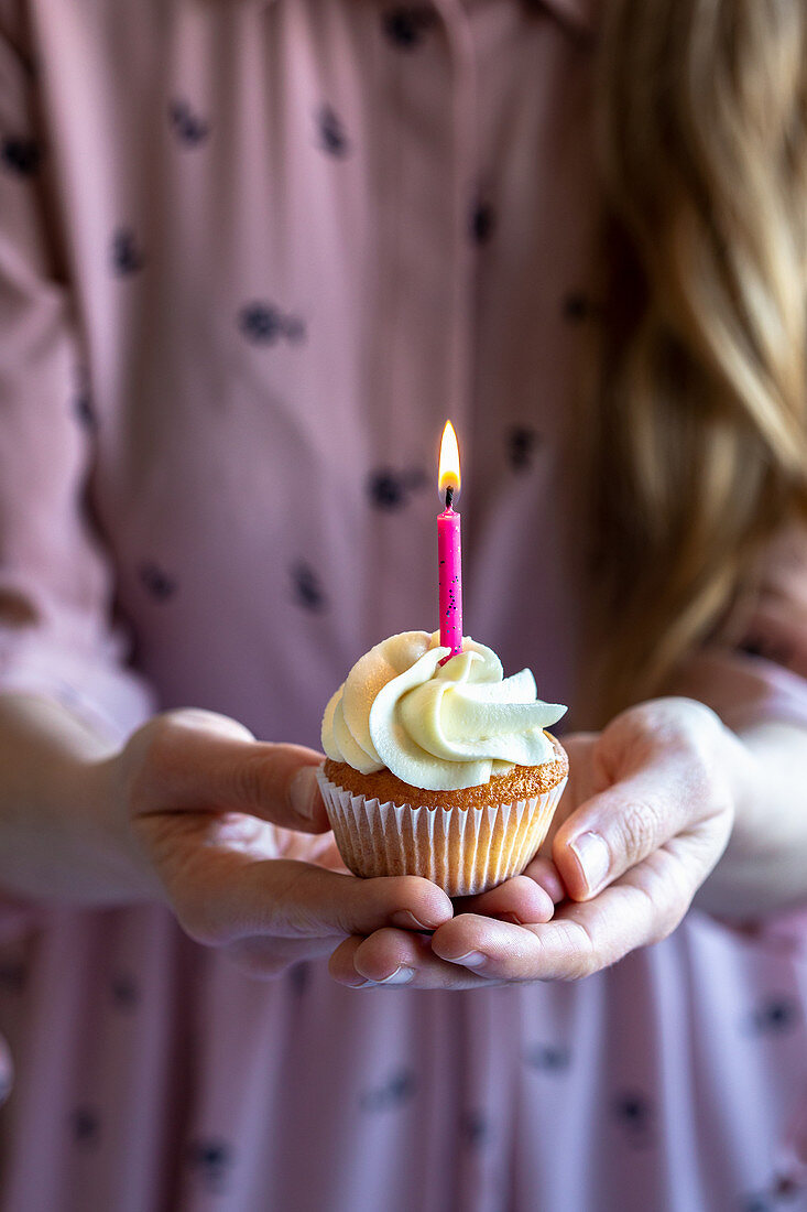Woman in pink dress and blond hair holding with two hands a cupcake with a pink candle burning