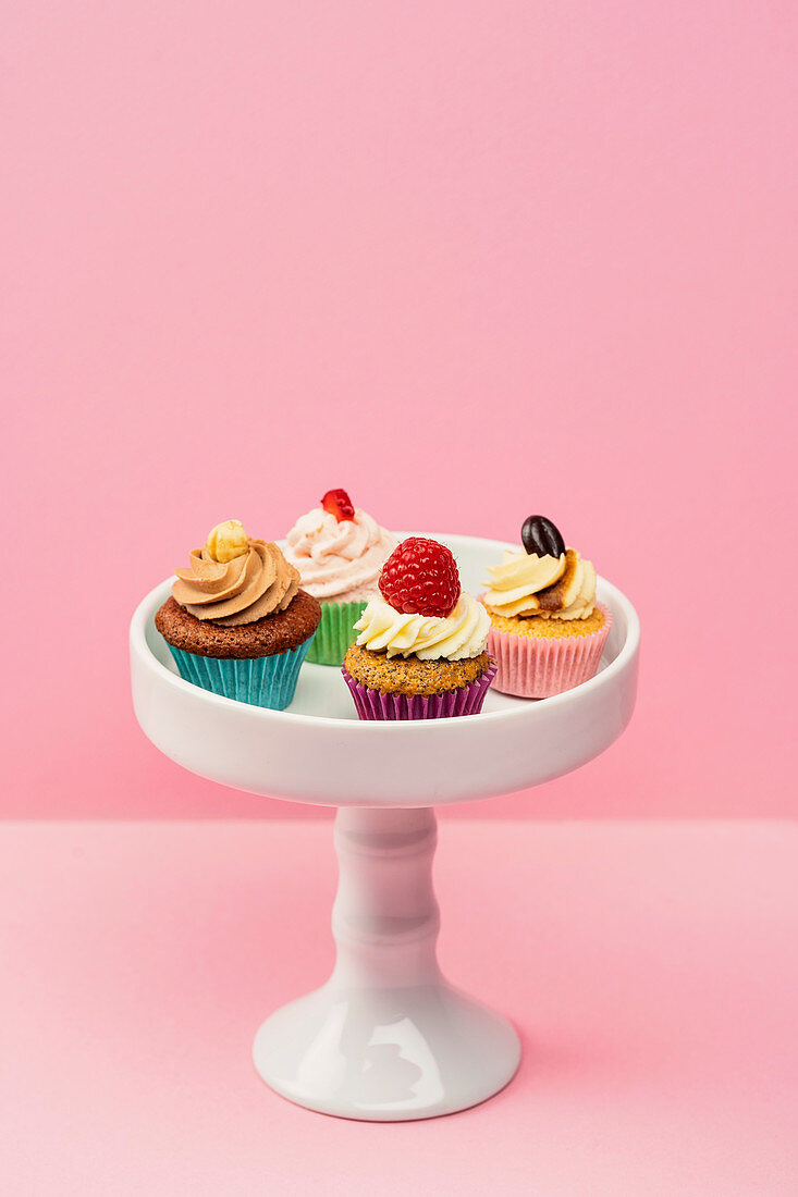 Verschiedene Mini-Cupcakes auf Gebäckständer vor rosa Hintergrund