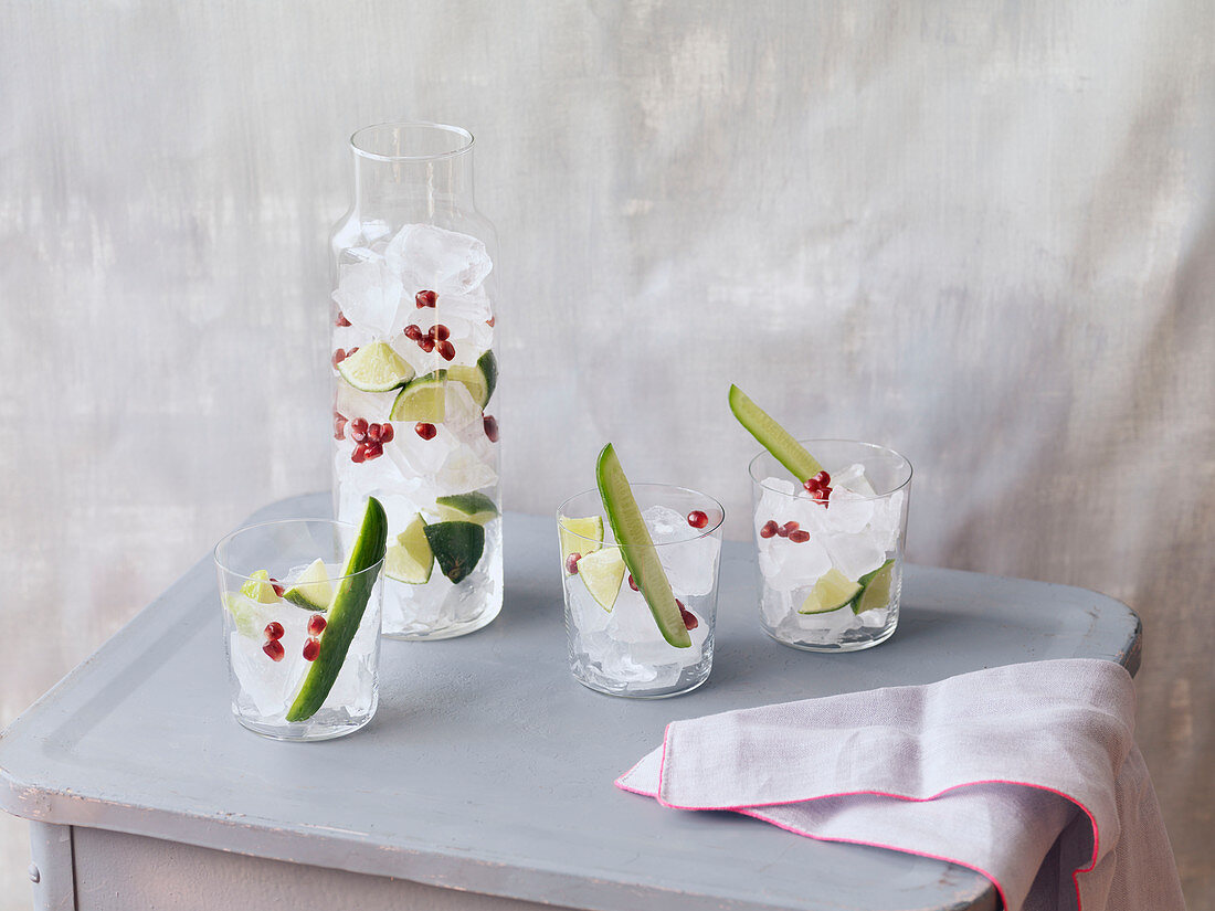 Three glasses and carafe of ice cubes, pomegranate and lime juice with cucumber garnish