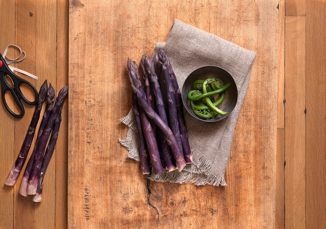 Purple Asapragus and Bowl of Fiddleheads with Scissors On Wood Surface