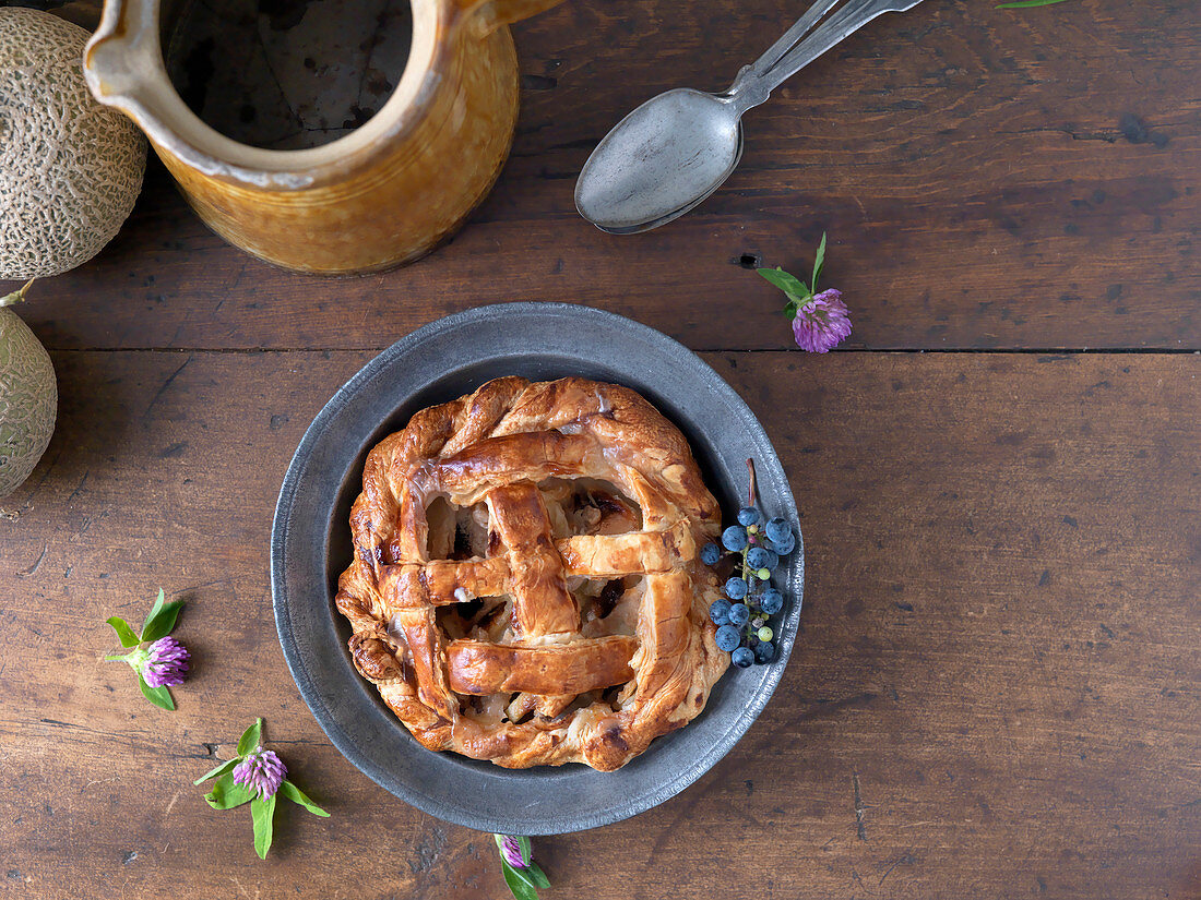 Rustic pie in pewter dish with antique spoon, jug, and melon on wooden surface