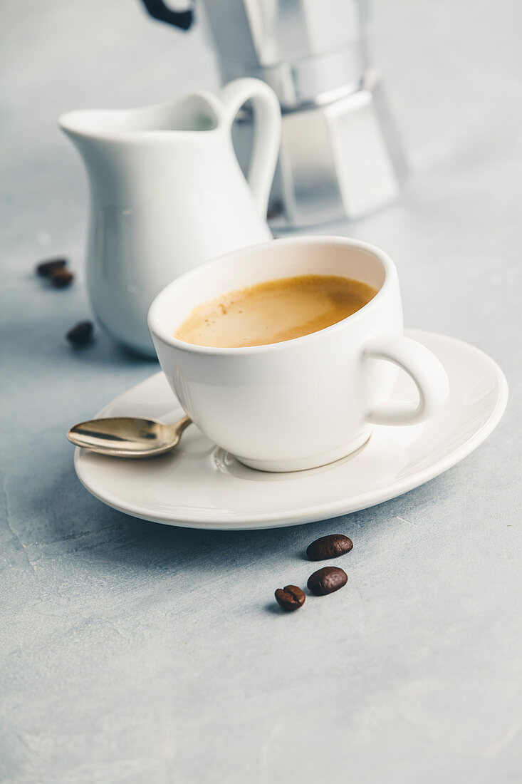 Eine Tasse Espresso mit Milchkännchen und Kaffeebereiter im Hintergrund
