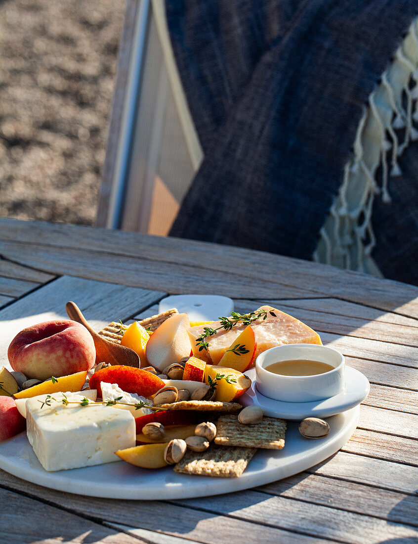 An outdoor table with a cheeseboard with feta, parmesan, crackers, pistachios, peaches, thyme and honey
