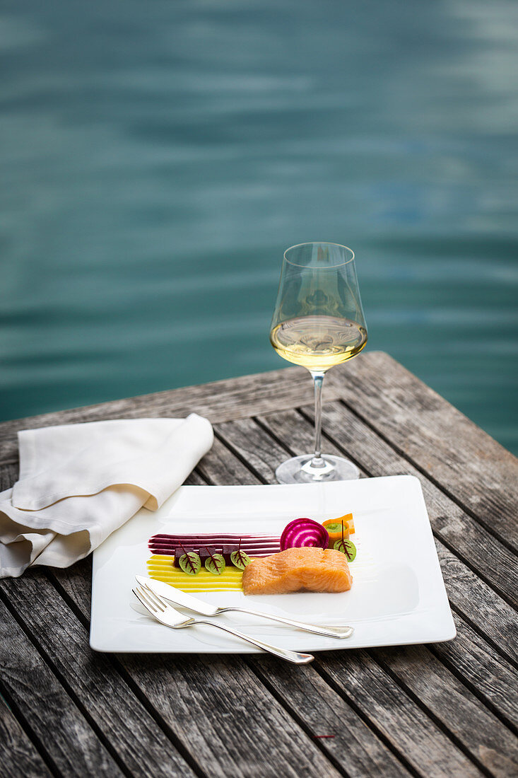 Saiblingsgericht und ein Glas Weißwein, serviert auf Holztisch mit Blick auf einen See