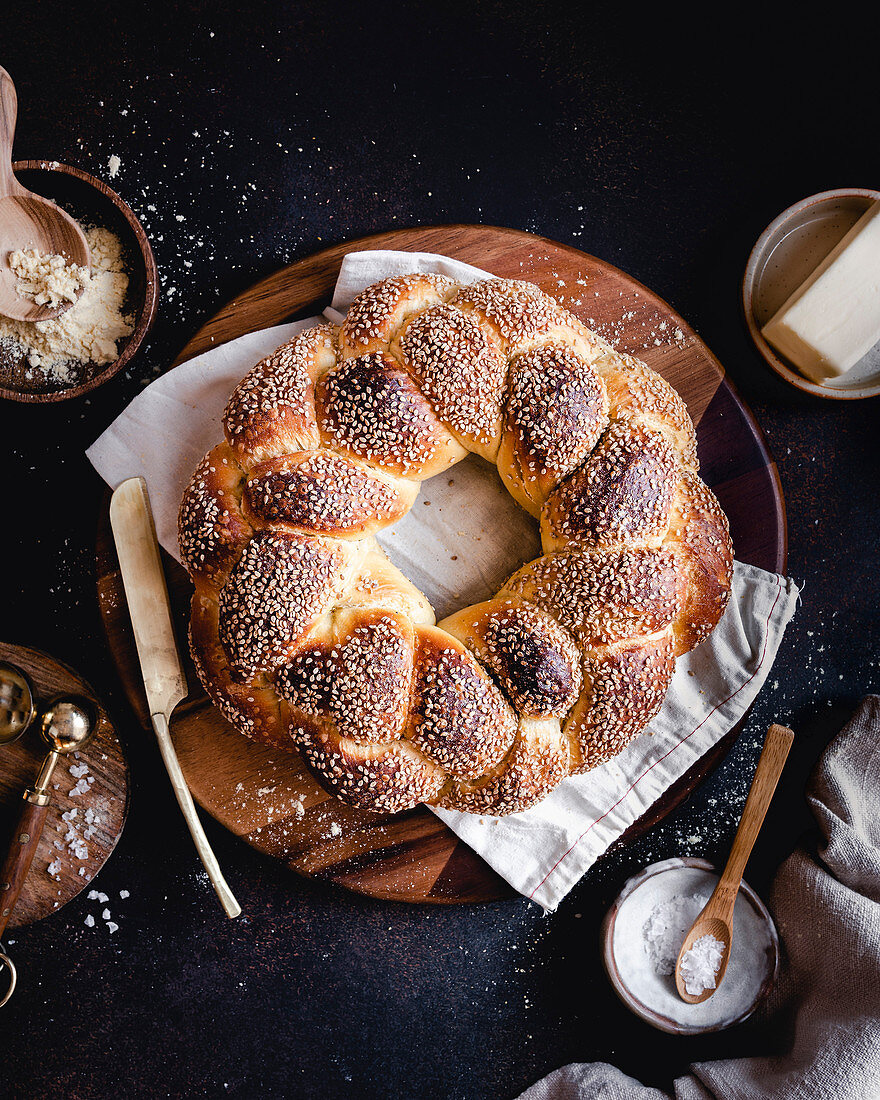 A sesame Challah bread wreath on a round wood board, with flour, seasalt and butter