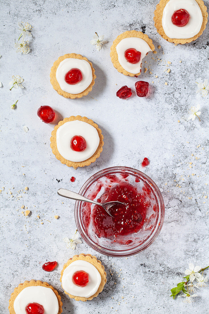 A line of jam filled shortbread biscuits topped with glace icing and cherries alongside a bowl of jam