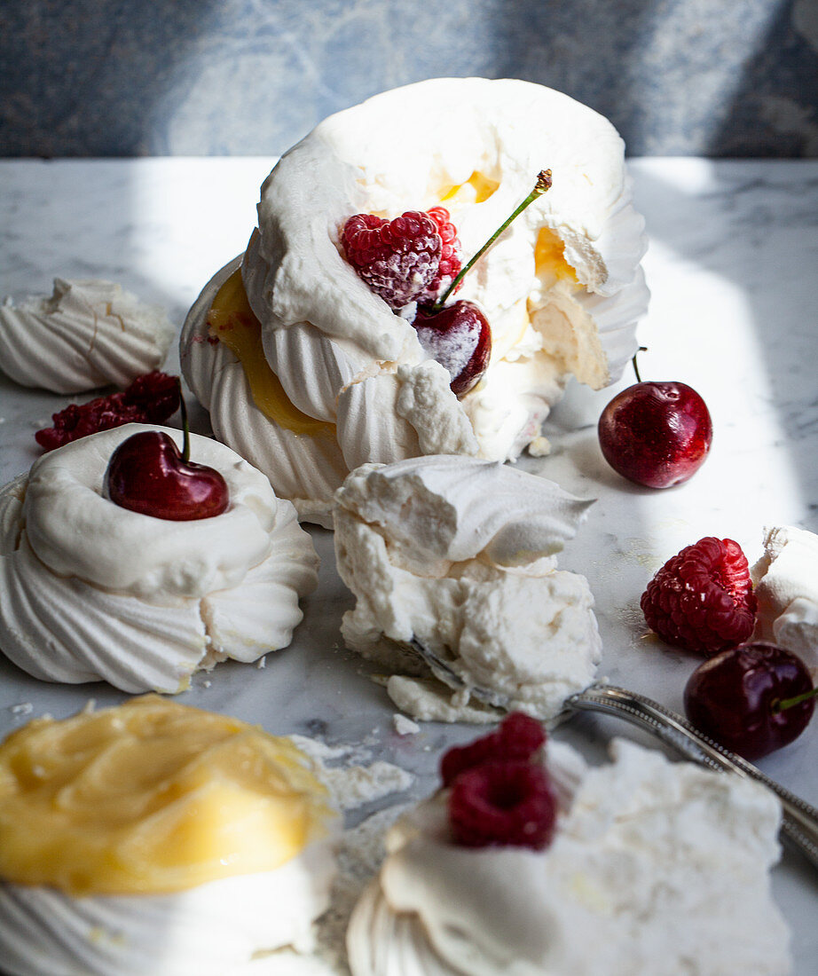 A tower of meringues topped with lemon curd, raspberries, whipped cream and cherries that has been toppled over