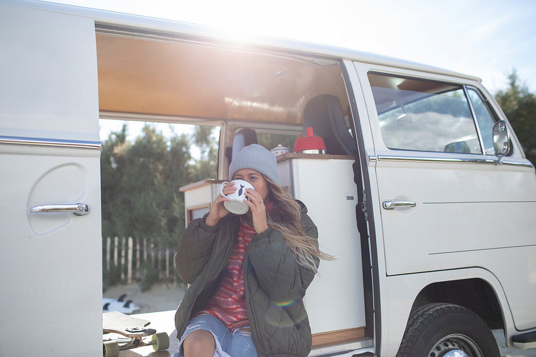 Young woman drinking coffee in sunny doorway of camper van