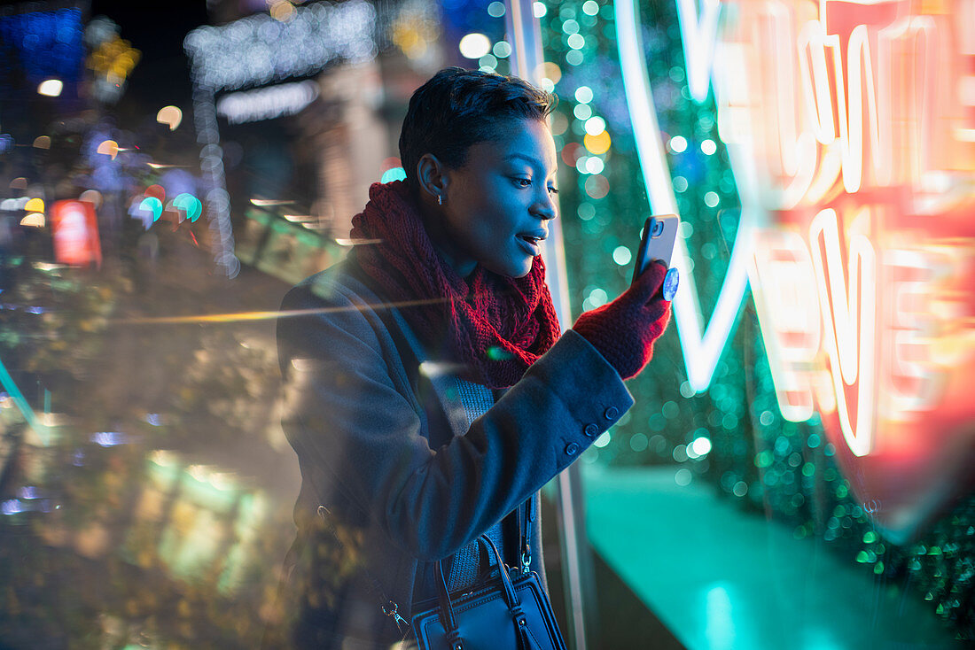 Young woman with smartphone at neon storefront sign at night