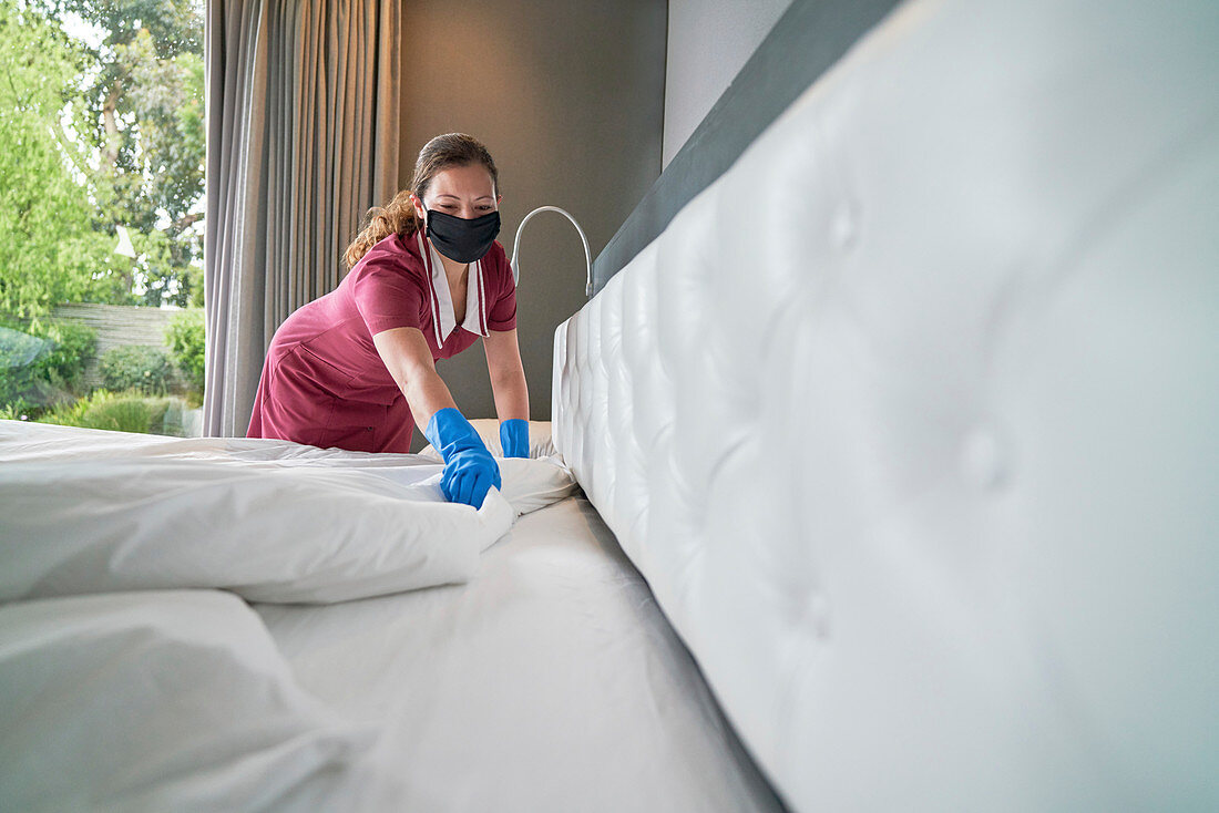 Female hotel maid in face mask making bed in hotel room