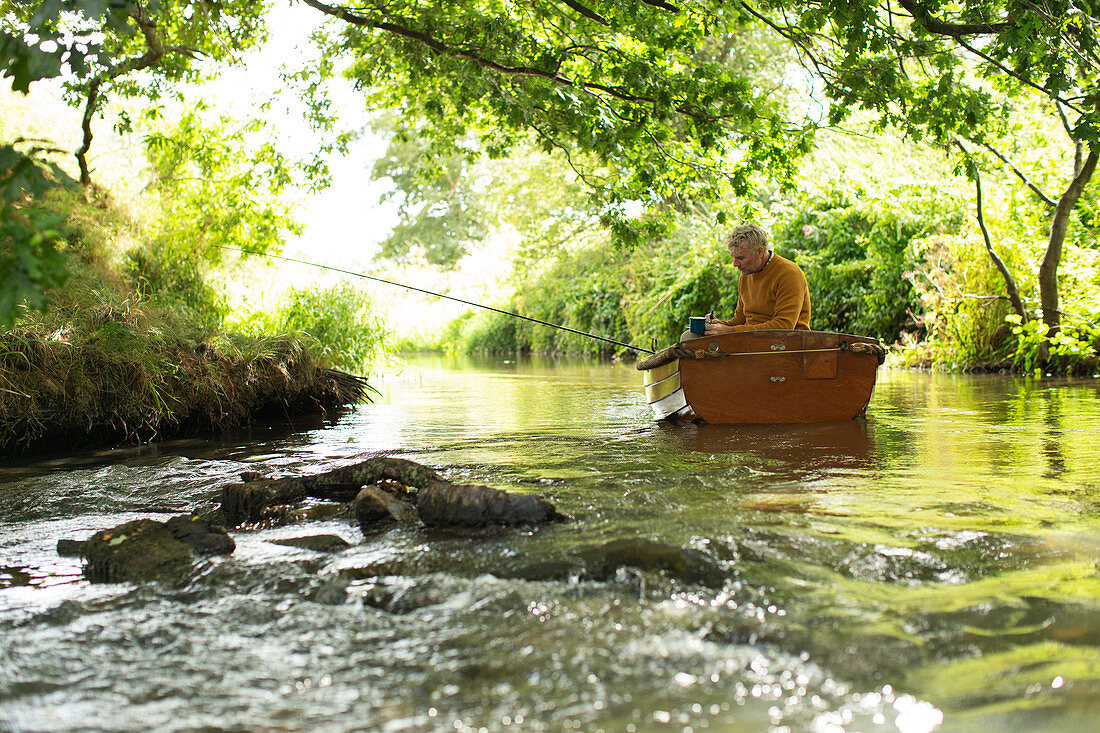 Man with coffee fly fishing and writing in boat on river