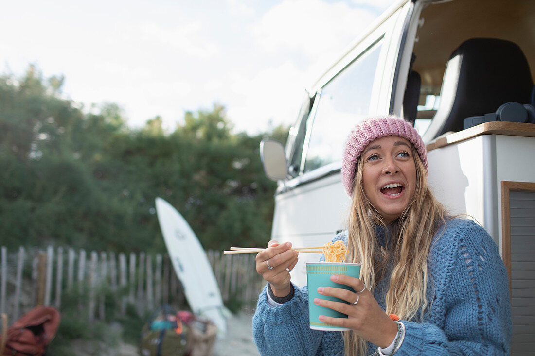 Happy woman eating instant noodles in camper van doorway
