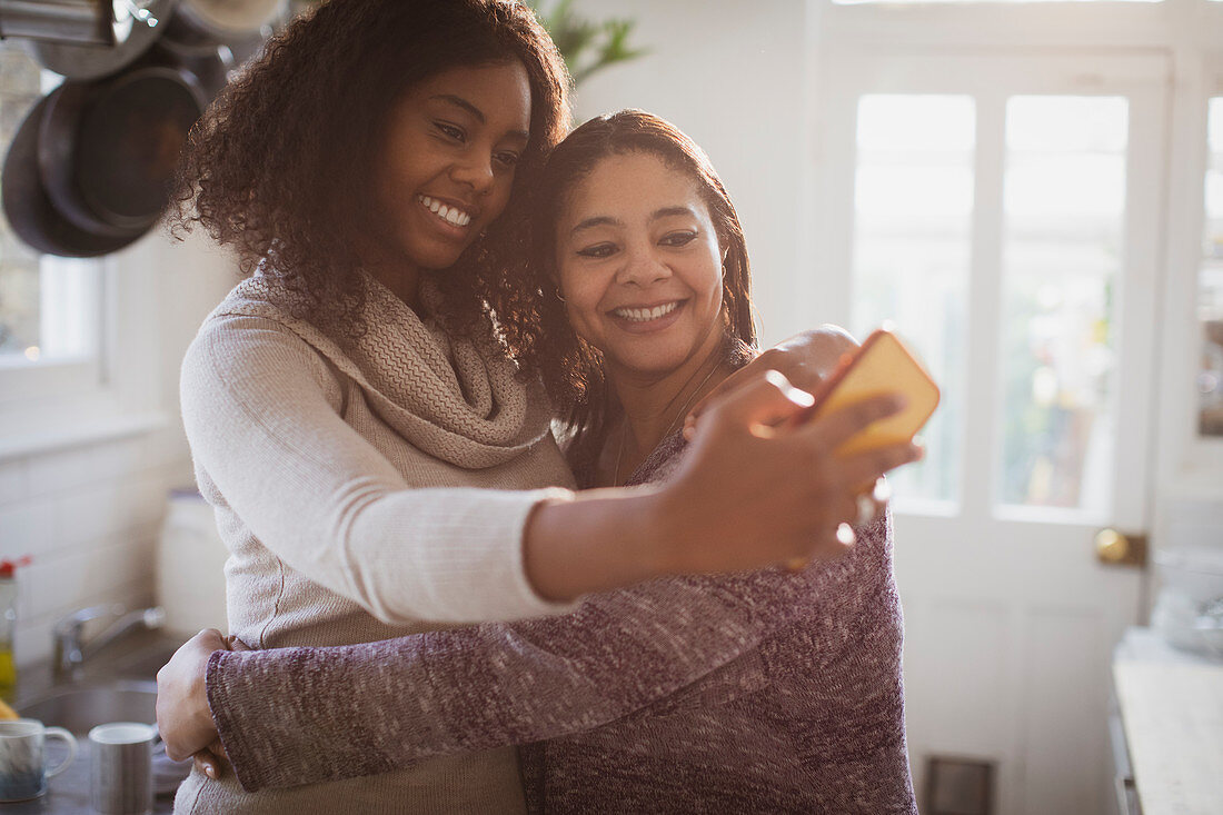 Mother and daughter hugging and taking selfie in kitchen