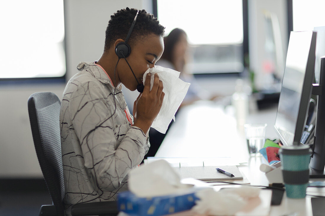 Woman in headset blowing nose into tissue at office desk