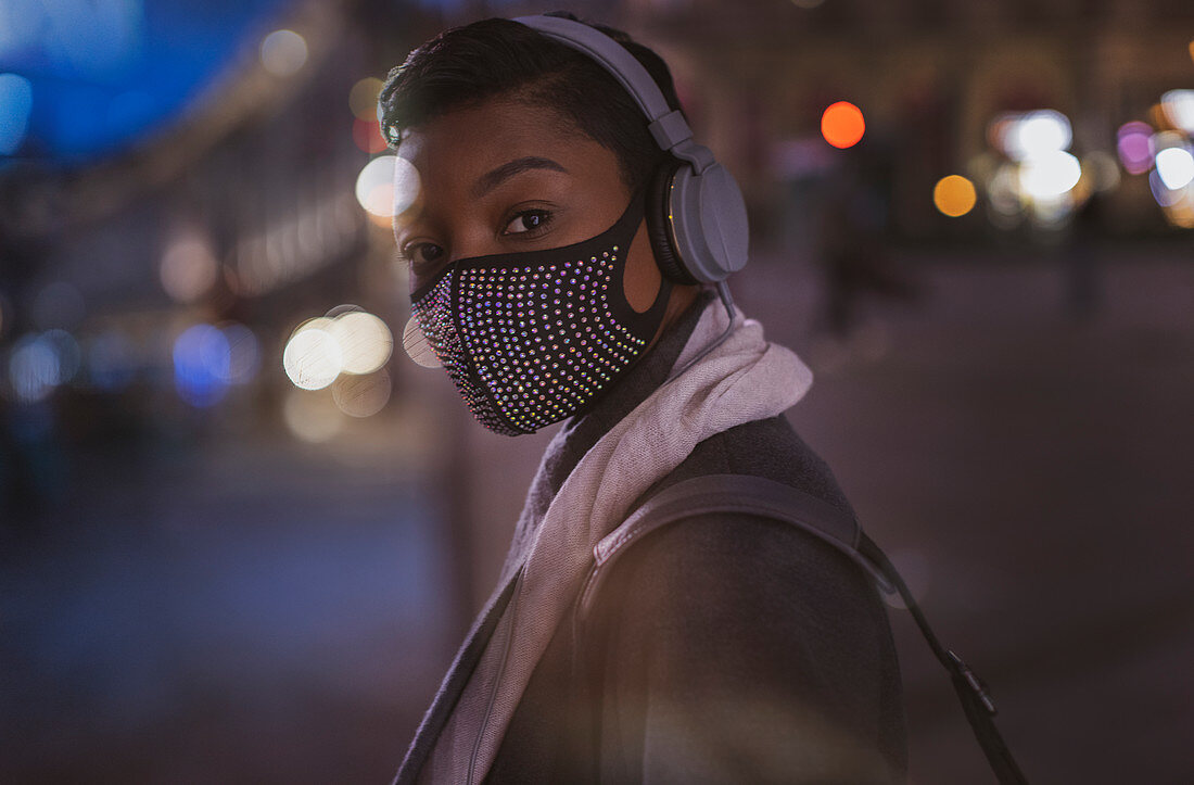 Young woman in facemask with headphones on street at night