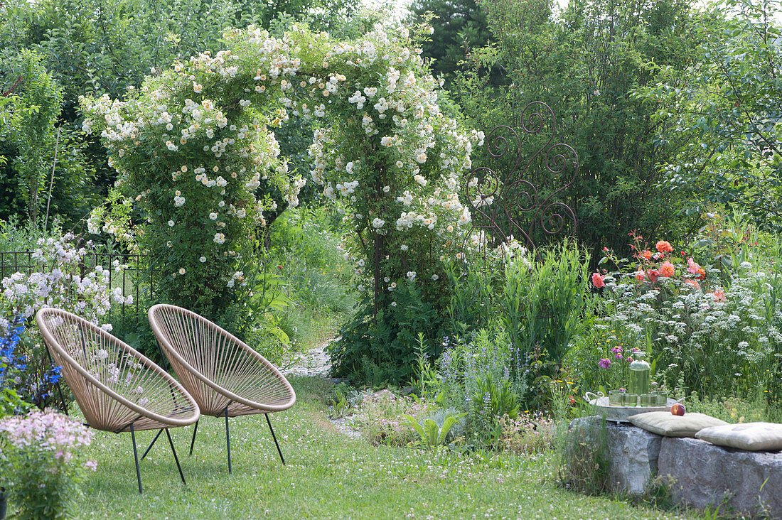 Rose arch with rambler rose 'Ghislaine de Feligonde', Acapulco chair on the lawn, cushion as seat on natural stones, decorative perennial support in the bed