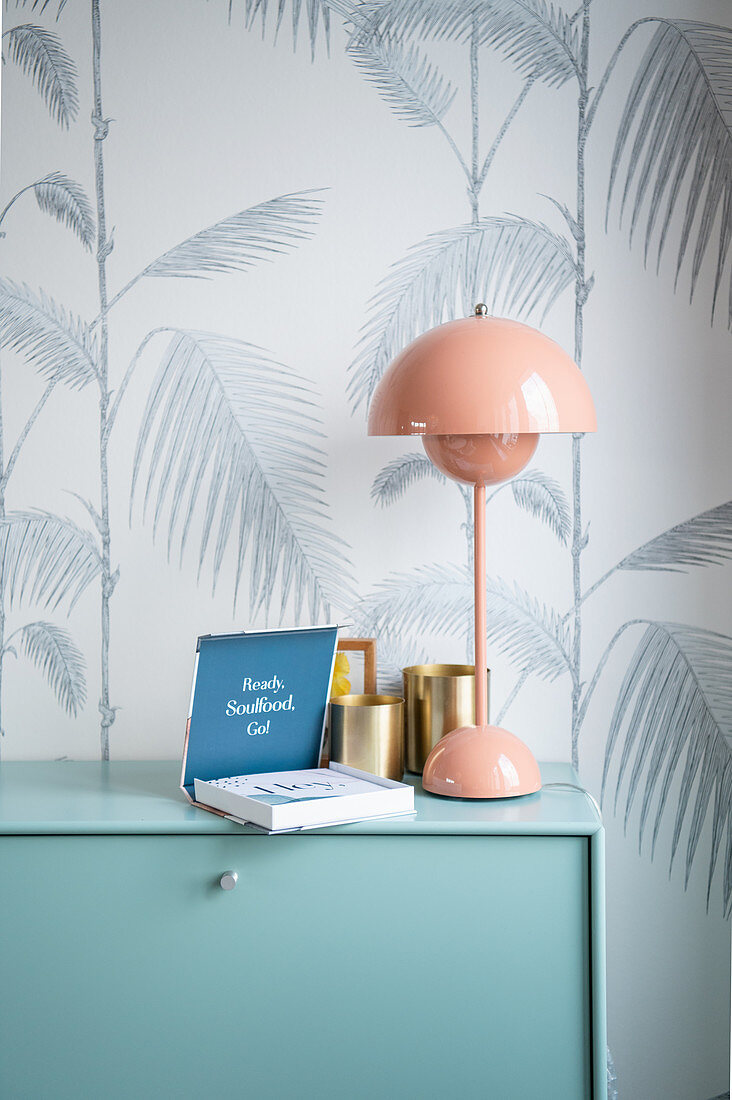 Table lamp, book and brass mug on a chest of drawers in front of wallpaper