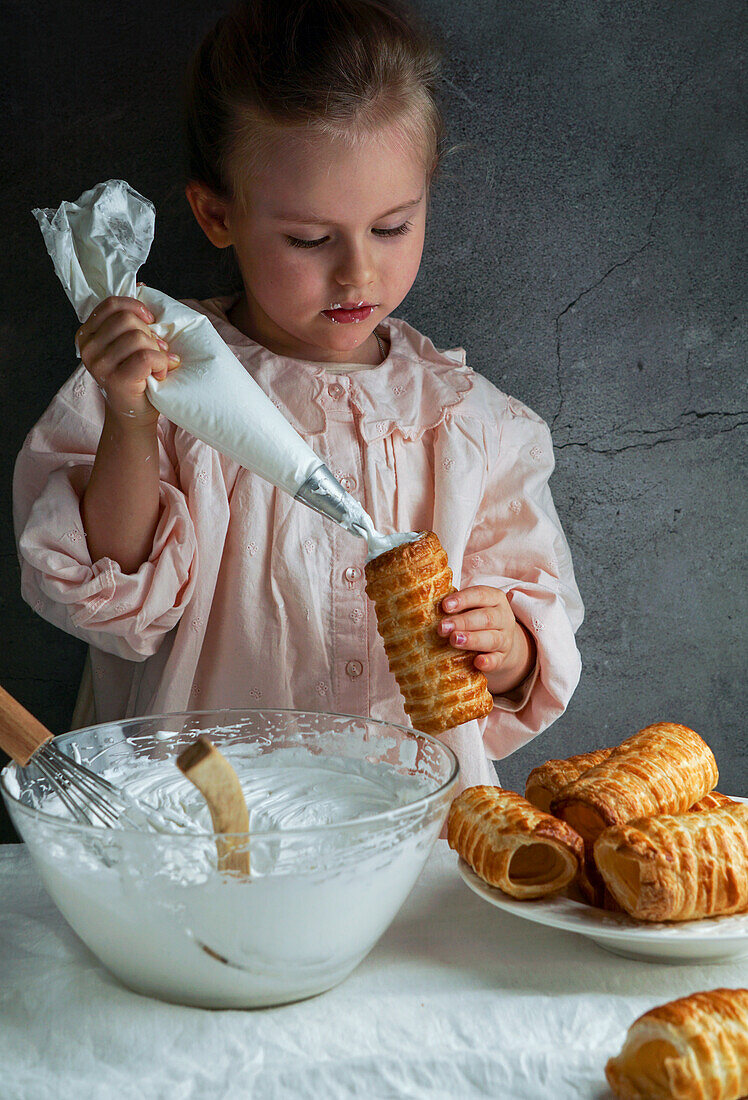 Little beautiful girl prepares puff rolls and fills them with cream