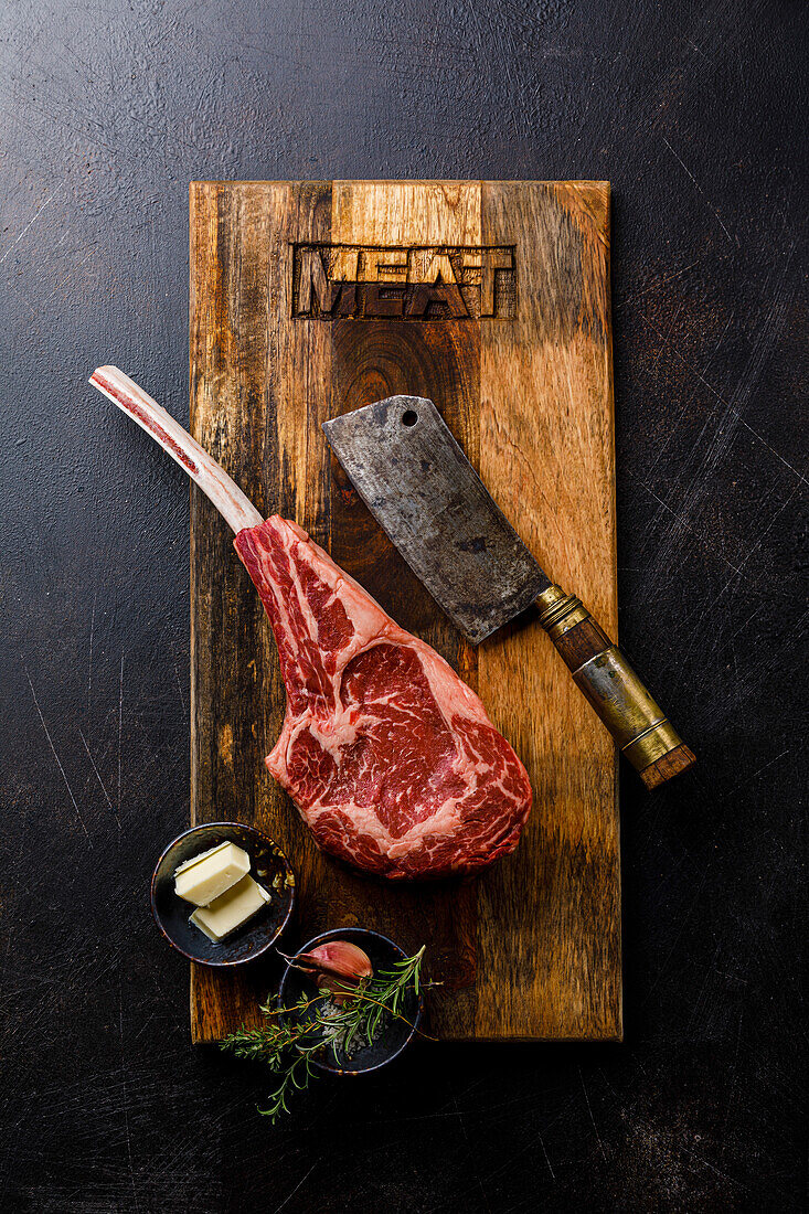 Raw fresh meat Tomahawk Steak, butcher cleaver and seasonings on wood board on dark background