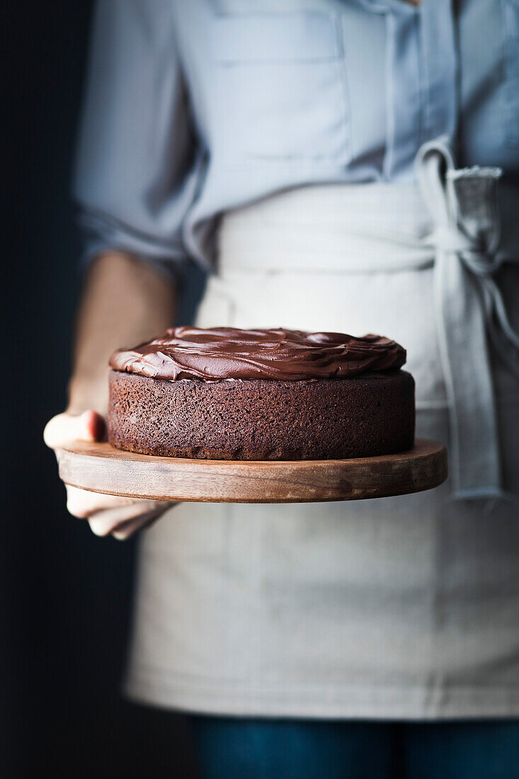 A person holding a chocolate cake on a wood cake board.