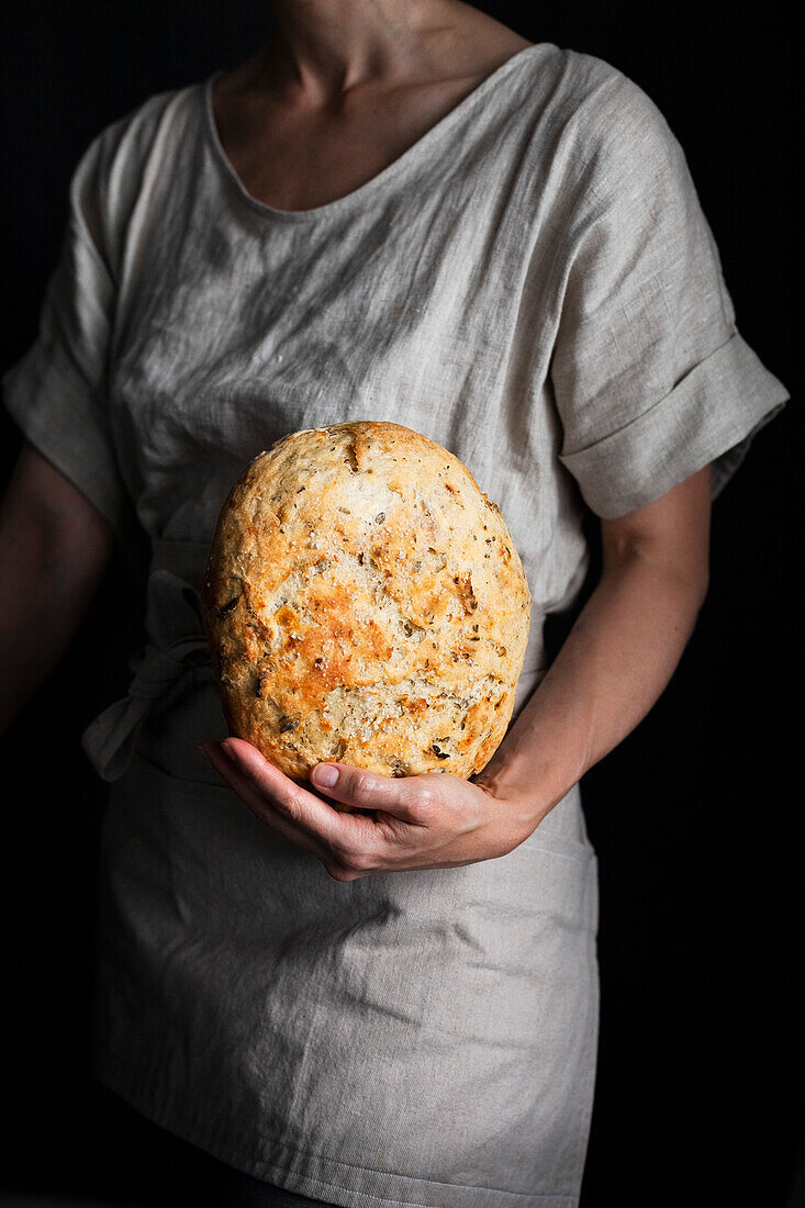 Person mit Leinenshirt und Schürze hält frisch gebackenen Brotlaib in der Hand
