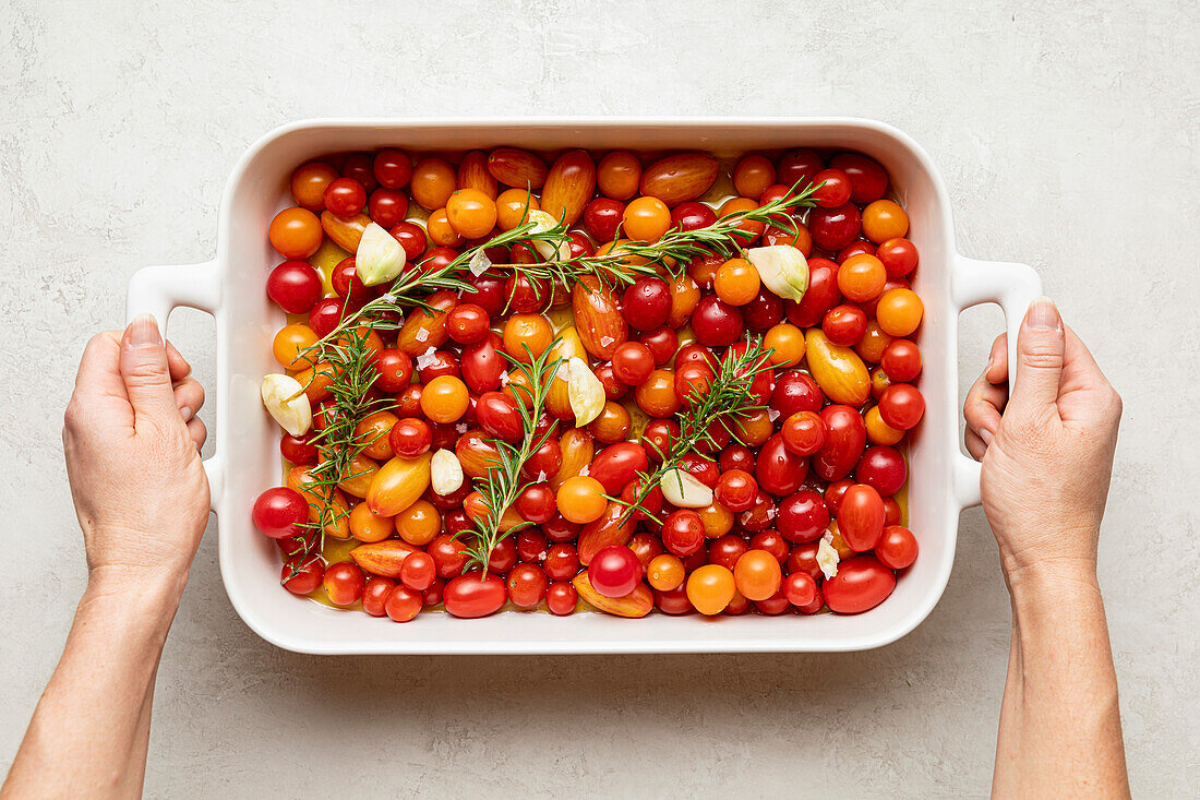 White baking dish filled with cherry tomatoes, garlic, rosemary, olive oil, and sea salt flakes