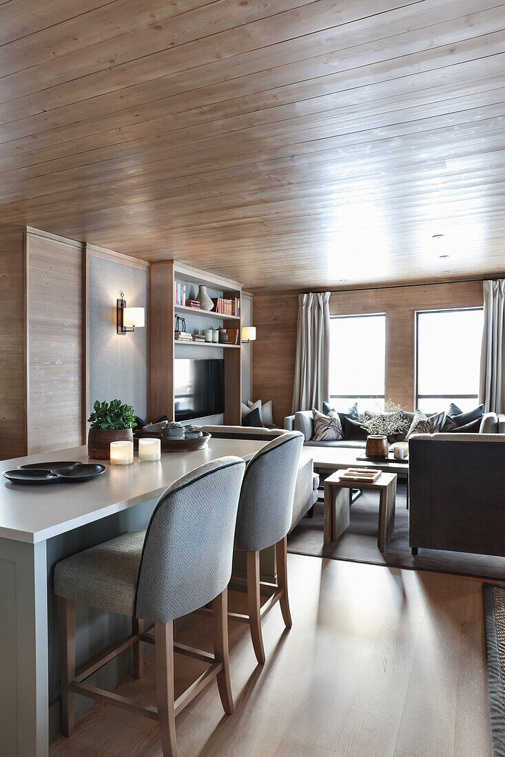 Kücheninsel mit Polsterstühlen, im Hintergrund Lounge in elegantem Wohnraum mit Holzverkleidung