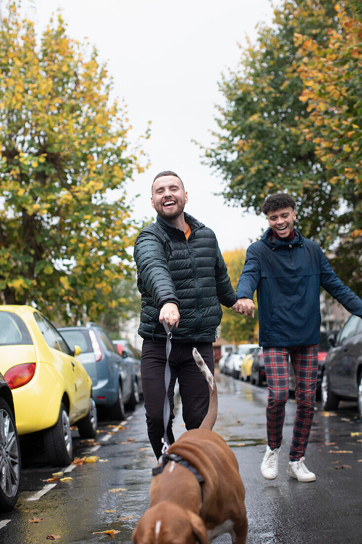 Happy gay male couple walking dog on wet autumn street