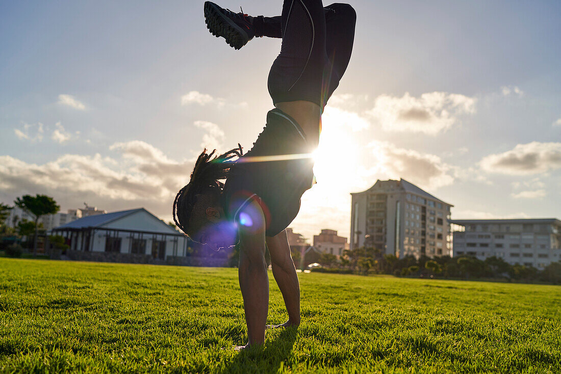 Athletic young man doing handstand in sunny park grass